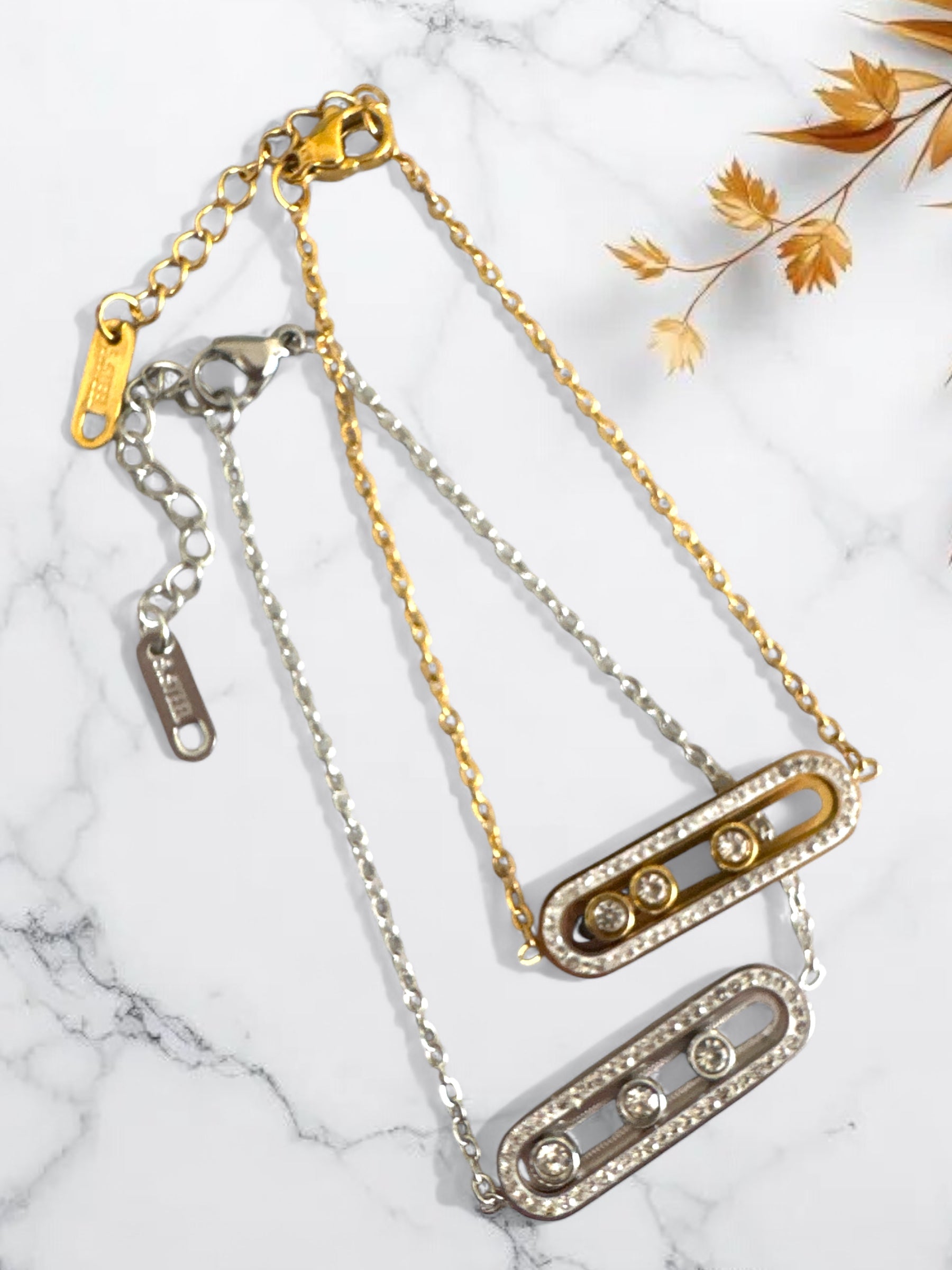 Δύο ατσάλινα βραχιόλια με τριπλά ζιργκόν, σε χρυσό και ασημί, τοποθετημένα σε φόντο μαρμάρου με φθινοπωρινά φύλλα