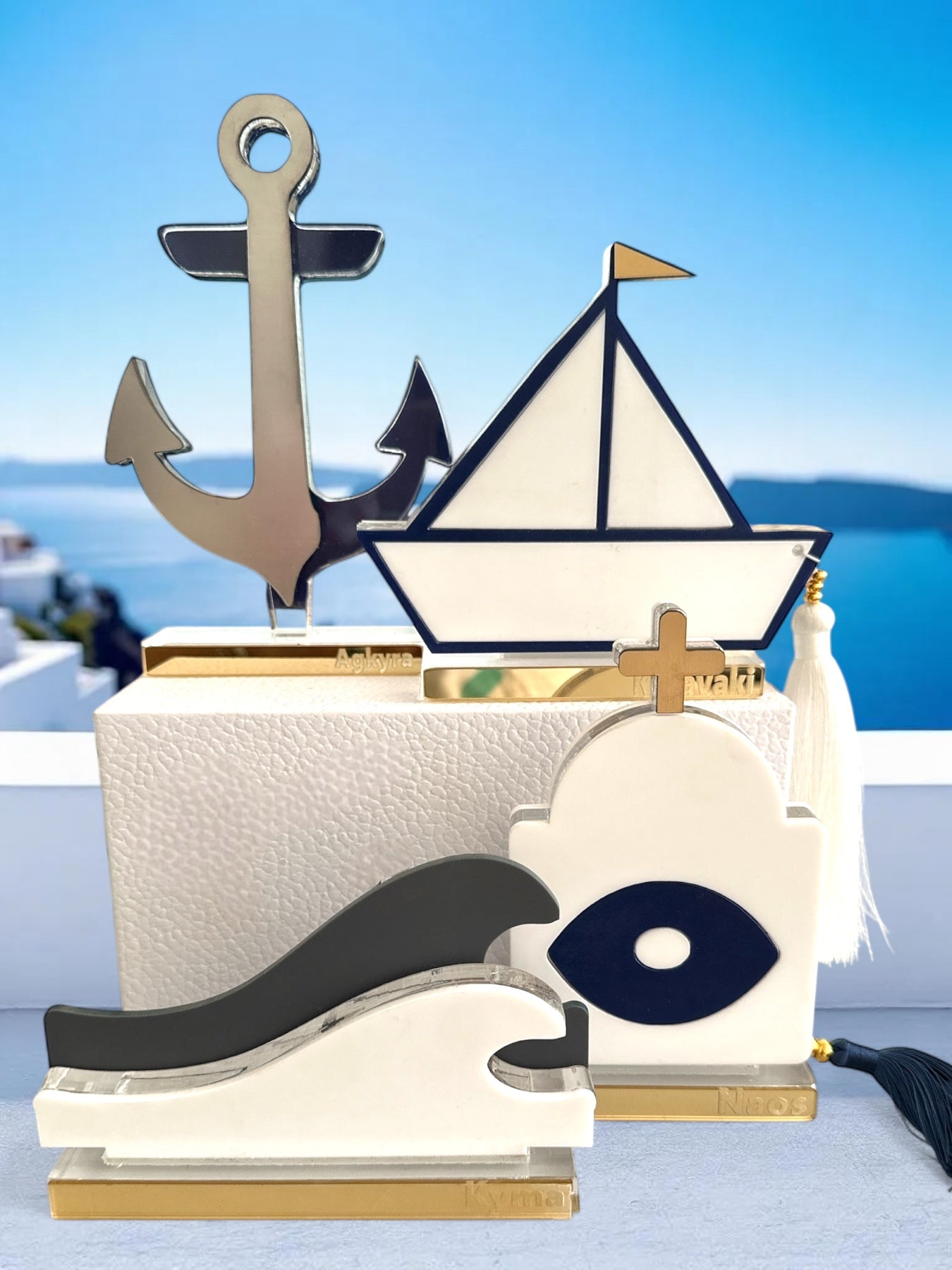 Σύγχρονα διακοσμητικά γούρια από πλεξιγκλάς σε κυκλαδίτικο φόντο: άγκυρα από ασημί καθρέφτη, καραβάκι με navy περίγραμμα και φούντα, κύμα σε λευκό-γκρι, και ναός με ματ μάτι – όλα σε διάφανη και χρυσή βάση.