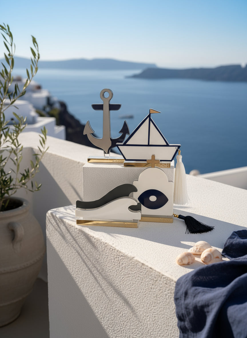 Σύγχρονα διακοσμητικά γούρια από πλεξιγκλάς σε κυκλαδίτικο φόντο: άγκυρα από χρυσό καθρέφτη, καραβάκι με navy περίγραμμα και φούντα, κύμα σε λευκό-γκρι, και ναός με ματ μάτι – όλα σε διάφανη και χρυσή βάση.