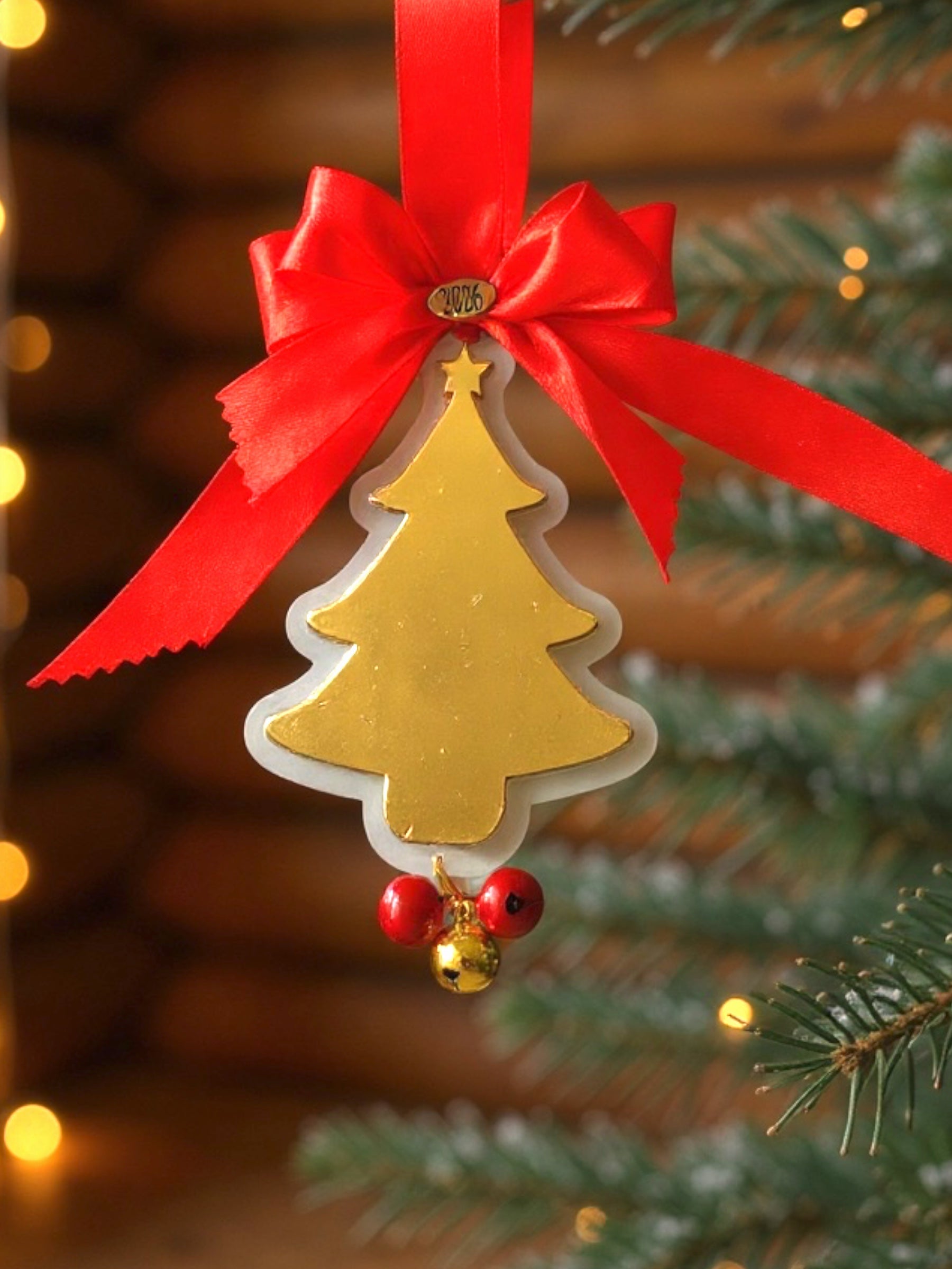 Decorative Christmas ornament with gold tree design, red ribbon, and small red berries on a blurred Christmas tree background.