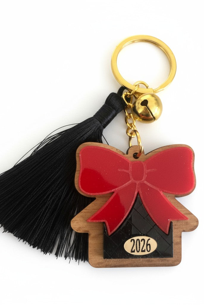 Χειροποίητο μπρελόκ γούρι 2026 με ξύλινο σπίτι, κόκκινο plexi φιόγκο, μαύρη φουντίτσα και χρυσό κουδουνάκι.