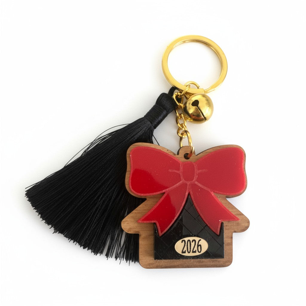 Χειροποίητο μπρελόκ γούρι 2026 με ξύλινο σπίτι, κόκκινο plexi φιόγκο, μαύρη φουντίτσα και χρυσό κουδουνάκι.
