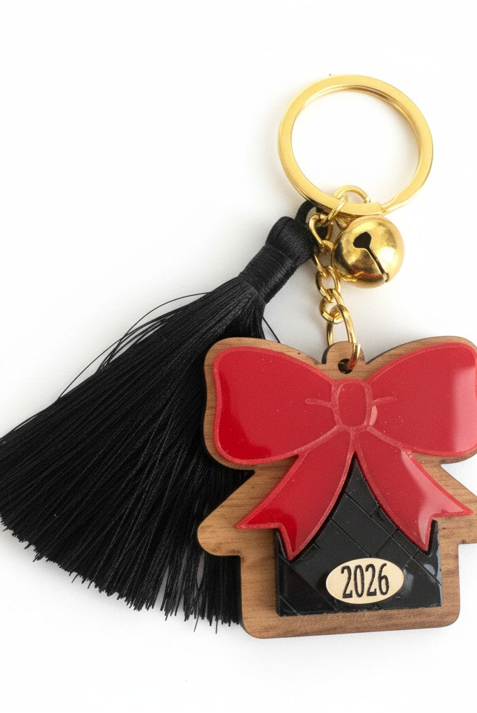 Χειροποίητο μπρελόκ γούρι 2026 με ξύλινο σπίτι, κόκκινο plexi φιόγκο, μαύρη φουντίτσα και χρυσό κουδουνάκι.