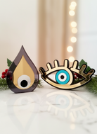 Επιτραπέζια γούρια από plexiglass σε σχέδιο σταγόνας και ματιού με βλέφαρα, σε χρυσό και τυρκουάζ mirror, τοποθετημένα σε festive σκηνικό με κλαδιά πεύκου και χριστουγεννιάτικα φωτάκια