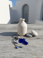 Μπρελόκ με μπλε ακρυλικό σταυρό και γκρι χάντρες, σε κυκλαδίτικη αυλή με λευκούς τοίχους και ξύλινη επιφάνεια.