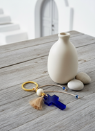 Μπρελόκ με μπλε ακρυλικό σταυρό και χρυσό κρίκο, τοποθετημένο σε ξύλινο τραπέζι δίπλα σε κεραμικό βάζο και λευκές πέτρες.