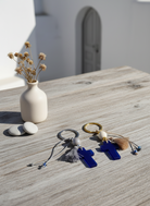 Δύο χειροποίητα μπρελόκ με μπλε σταυρό σε κυκλαδίτικο σκηνικό, πάνω σε ξύλινο τραπέζι με κεραμικό βάζο και λευκές πέτρες.