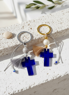 Δύο μπρελόκ με μπλε ακρυλικό σταυρό, φουντάκια και κορδόνια με ματάκια σε λήψη επάνω σε λευκή κυκλαδίτικη επιφάνεια.