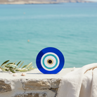 Γούρι μάτι από μπλε plexiglass πάνω σε πέτρινο τοίχο με φόντο τη θάλασσα και διακοσμητικό κλαδί ελιάς