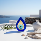 Plexiglass teardrop evil eye decor Milos on white terrace with Aegean sea background.